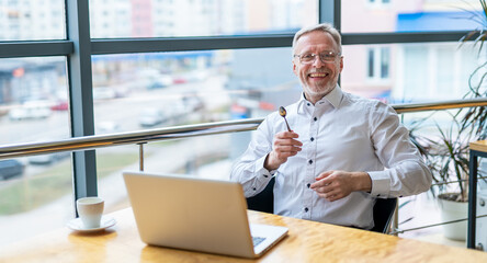 Smiling middle aged businessman in white shirt with a laptop. Man sitting near the window working with documents.