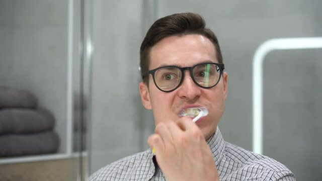 Young Caucasian man with toothbrush cleaning teeth, looking mirror in bathroom close up. Handsome guy in plaid shirt, black glasses brushing his teeth in morning before going to work. Stomatology care
