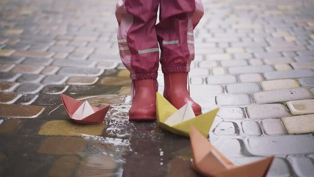 Feet of little girl in rubber boots stomping in puddle with paper boats around. Unrecognizable happy kid having fun playing outdoors on rainy day. Childhood, leisure, fun.