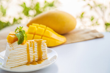 A mango crepe cake sliced on a white plate decorated with a piece of mango fruits and peppermint on a wite table with mango fruit and green nature background.