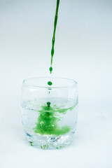Green paint is poured into a transparent glass with clear water in the movement and paints clear water green on a white background.