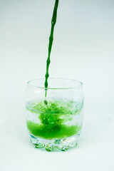 Green paint is poured into a transparent glass with clear water in the movement and paints clear water green on a white background.