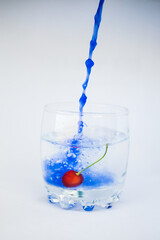 In a transparent glass with clean water, with cherries at the bottom, blue paint is poured in a dynamic movement and paints the water blue on a white background.