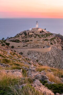 soft dreamy purple pink morning dawn sunrise vertical pic of cap de formentor light house on dry rocky sand cliff with yellow dry grass wavy road with sea ocean on background. Mallorca Majorca Spain