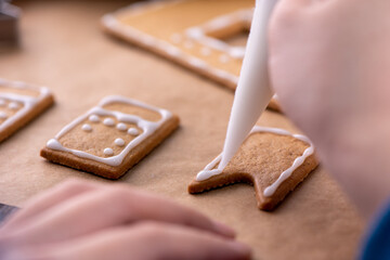 Young woman is decorating Christmas Gingerbread House cookies biscuit at home with frosting topping in icing bag, close up, lifestyle.