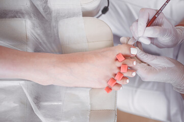 Pedicurist doing white nail polish on client legs using shellac lamp. Professional medical pedicure procedure. Foot treatment in SPA salon. Podiatry clinic. Beautician doctor hands in white gloves.