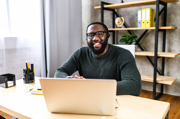 Attractive African-American guy uses a handsfree headset and laptop to talk online at his workplace, black confident man in glasses sits at the office desk, looks at camera with pleasant smile