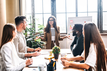 A team of young and successful employees listen to progress report from female employee at a morning meeting. Multiracial team in the office