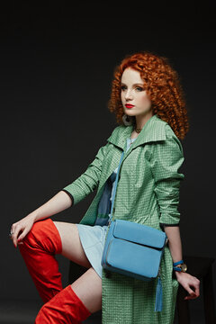 girl with red curly hair, white skin, a green cloak and high orange boots, dark background, blue bag,