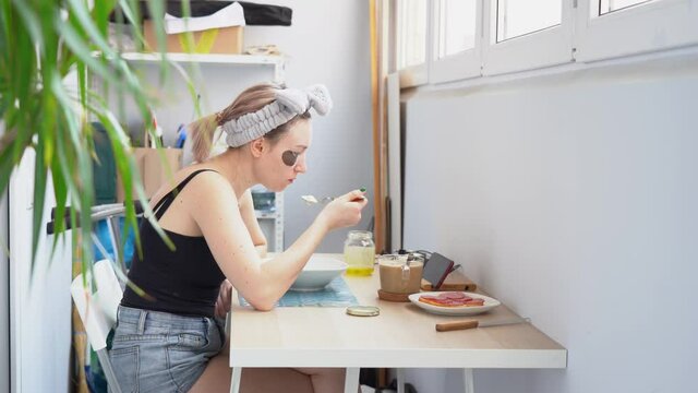 One young attractive blonde woman in black T-shirt, denim shorts wear hair band, sits at table, eats hot porridge, watches video on smartphone on white balcony at home. Morning breakfast. Side view.