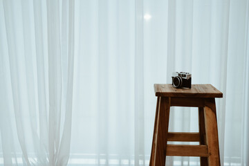 camera on wooden stool chair beside see through window curtain