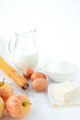 Jug with milk, eggs, flour, rolling pin, apples, butter and a whisk on a white table. The recipe for making apple pie.