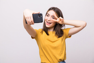 Portrait of a young attractive woman with peace gesture making selfie photo on smartphone isolated on a white background