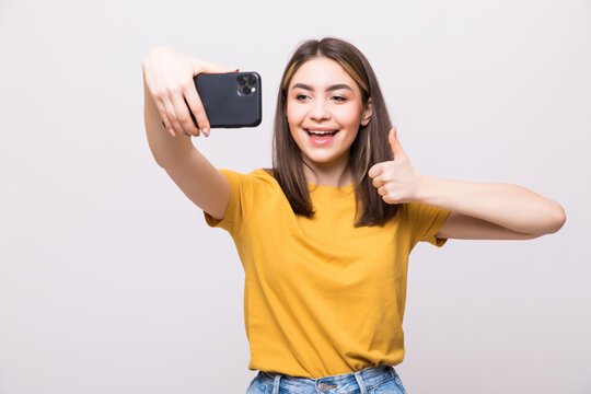 Portrait of a young attractive woman with thumbs up making selfie photo on smartphone isolated on a white background