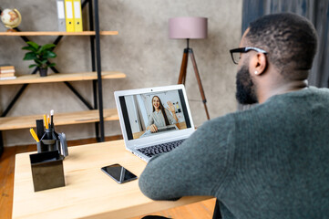 Back view African-American guy talks with female friend or coworker via video call, he sits at the office desk and using laptop for video meeting, a young woman on the screen