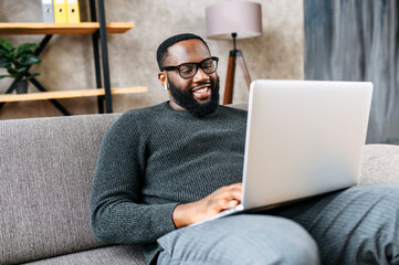Cheerful African-American guy sits on sofa in modern living room alone and using trendy laptop for net searching, web surfing or online learning