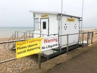 A sign on seaside railings warns that no Lifeguard is present and reminds people to observe social distancing.A life guard station is visible with beach and sea in background.Image