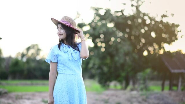 An Asian woman is wearing a blue dress and is wearing a hat standing at the rice field at sunset.
