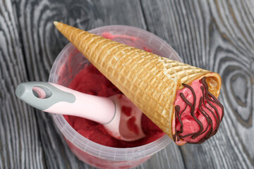 Strawberry ice cream in a waffle cone. Garnished with Chocolate. Near the container with ice cream and a spoon for it. On pine boards painted in black and white.