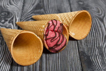 Strawberry ice cream in a waffle cone. Garnished with Chocolate. Nearby are empty waffle cones without ice cream. On pine boards painted in black and white.