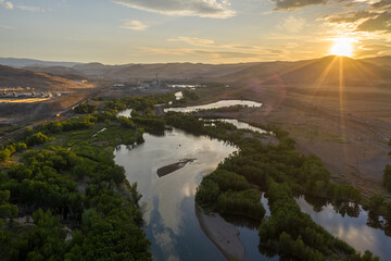 A tree lined river with an island through a desert mountain range, a railroad and a major highway, and the sun setting over the horizon