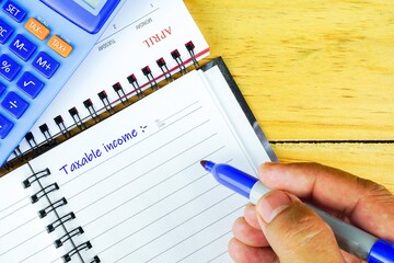Conceptual image of tax return to tax authority in April. Fingers holding pen writing on blank note book. Calculator visible. Selective focus on beads. Wooden background.
