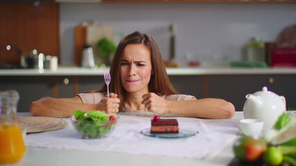 Obraz na płótnie Canvas Woman choosing cake instead salad on kitchen. Lady eating cake on kitchen