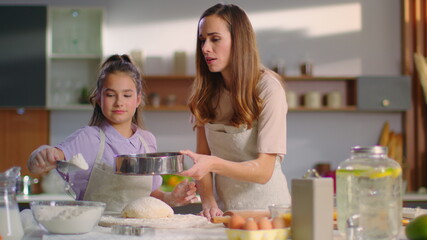 Woman teaching daughter to sieve flour on dough at kitchen