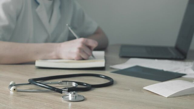 Doctor therapist working with laptop computer and writing on paperwork and looking X-ray of lungs, fluorography, roentgen in hospital. Woman in medical gown, stethoscope in consulting room.