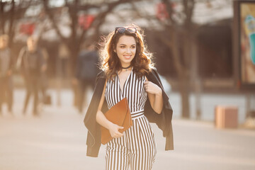 Fototapeta premium Beautiful brunette woman in stylish spring look, wearing in striped suit and black jacket, holding red handbag, Smiling girl with make up after beauty salon walking at street, posing at camera.Sunset.