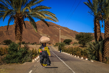 Eine marrokanische Frau geht auf einer Landstrasse und trägt auf dem Kopf einen Sack mit Getreide