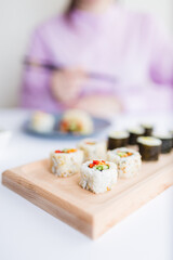 Sushi rolls on wooden plate in front of woman using chopsticks, japanese meal concept.