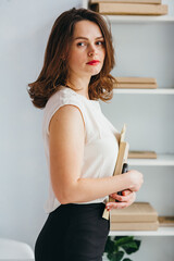 A girl in a white blouse stands with her phone and books in her hands in the office. She has brown...