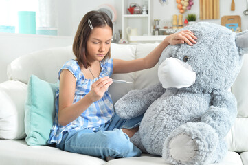 Cute girl with thermometer playing with toy teddy bear in facial mask