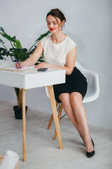 An elegant girl in a white shirt and black skirt sits at a white table. She thought. The girl sits in an office decorated in white and beige colors. 