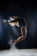 Fototapeta na wymiar Beautiful plump blonde girl wearing a black gymnastic bodysuit covered with clouds of the flying white powder jumps dancing sitting on a dark. Artistic conceptual and advertising photo.