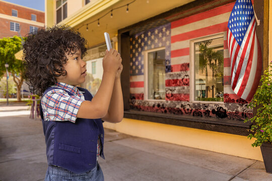 Curly hair boy takes a photo of an American flag on the building. The building has a framed painting of the American flag with outlined flower overlay with windows in the middle.