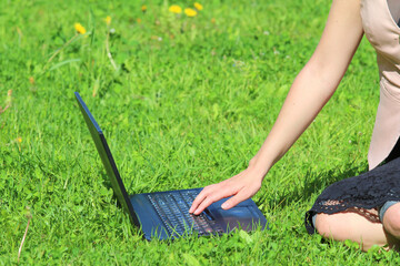 A beautiful young white girl in a pink jacket and black skirt and with long hair sitting on green grass, on the lawn and working behind a black laptop