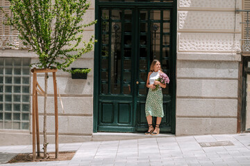 beautiful woman with bouquet of flowers waits in the doorway of a house. Pretty young girl with a portrait on the street.