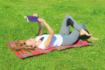 A beautiful young white girl in a white T-shirt and with long hair lies on a red plaid, on green grass, on the lawn and read book