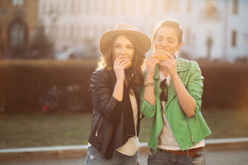 Positivity girls eating fast food, hamburgers, having dinner together, smiling at camera and posing. Beautiful couple of stylish girlfriends in leather jackets and hat, eating junk and unhealthy food.