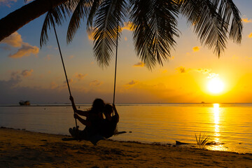 Silhouetted couple in love walks on the beach during sunset. Riding on a swing tied to a palm tree and watching the sun go down into the ocean