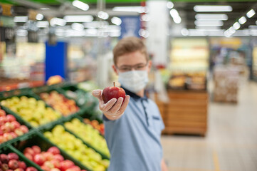 A teenager in a supermarket holding an apple. He's wearing a mask and gloves.