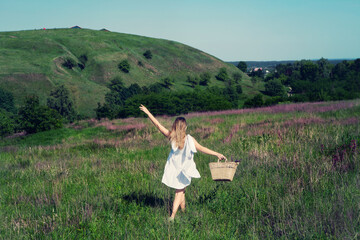 Beautiful girl in white dress with straw bag and wildflowers runs in meadow