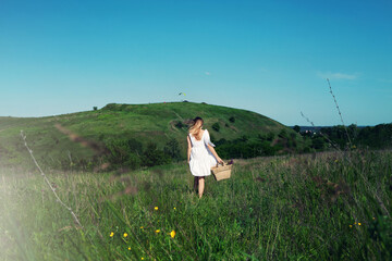 Beautiful girl in white dress with straw bag and wildflowers runs in meadow