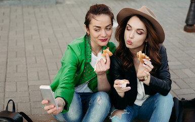 Positive and stylish girls eating hamburgers making duck face and taking self portrait at smart phone.Beautiful two women stylish wearing in hat and leather jacket, have dinner at street by fast food.