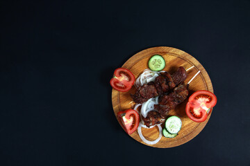 shashlick and cut vegetables on wooden cutting board on black background flat lay