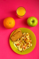 diet bread and granola on a plate, orange, apple and a glass of juice on a pink background.