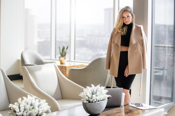 Fashionable beautiful classy, high class woman in coat in hotel lobby with windows showing city...
