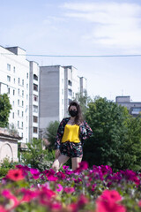Fototapeta na wymiar Attractive young brunette woman in yellow t-shirt and flowery shorts costume. In medical black face mask. modern reality. near bushes with flowers. covid-19 concept. copy space. High quality photo